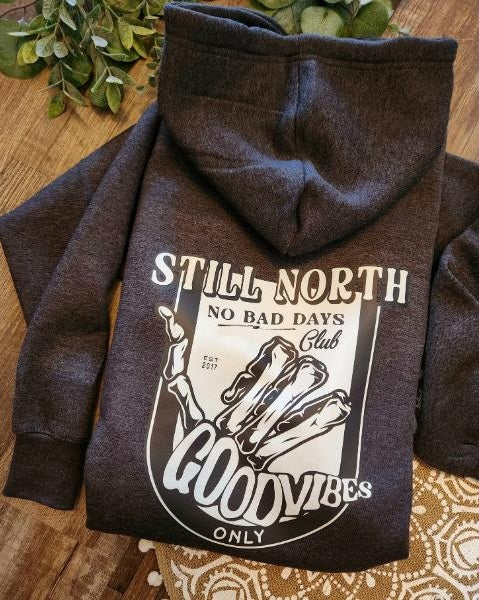 grey hoodie with 'Still North' and 'Good Vibes Only' text on a wooden surface.