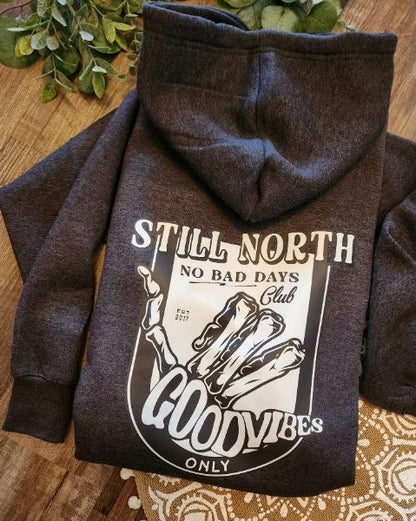 grey hoodie with 'Still North' and 'Good Vibes Only' text on a wooden surface.