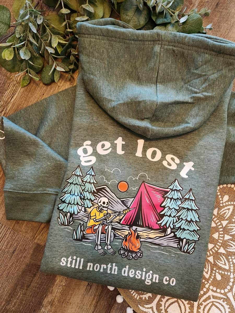 Green hoodie with 'get lost' and camping design on a wooden surface with greenery.