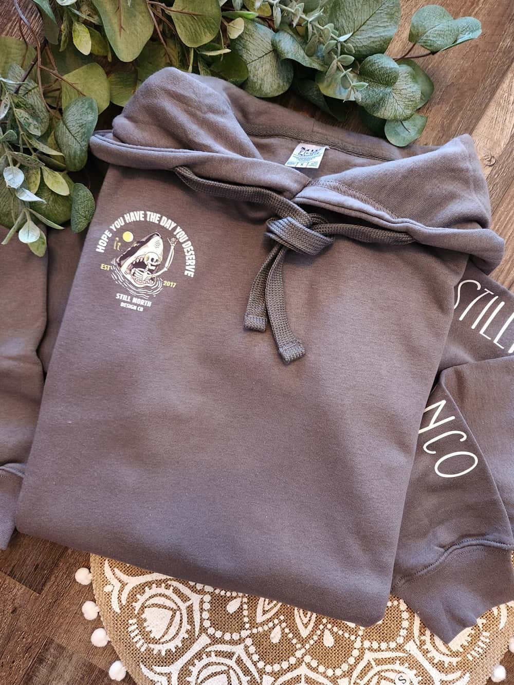 Folded brown hoodie with a logo on a wooden surface with greenery