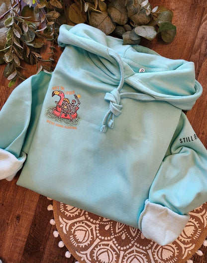 Light blue hoodie with a graphic design on a wooden surface with plants in the background