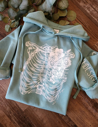 Light blue hoodie with a white skeleton and floral design on a wooden surface.