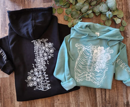 Two hoodies, one black and one teal, with floral and skeleton designs on a wooden surface.