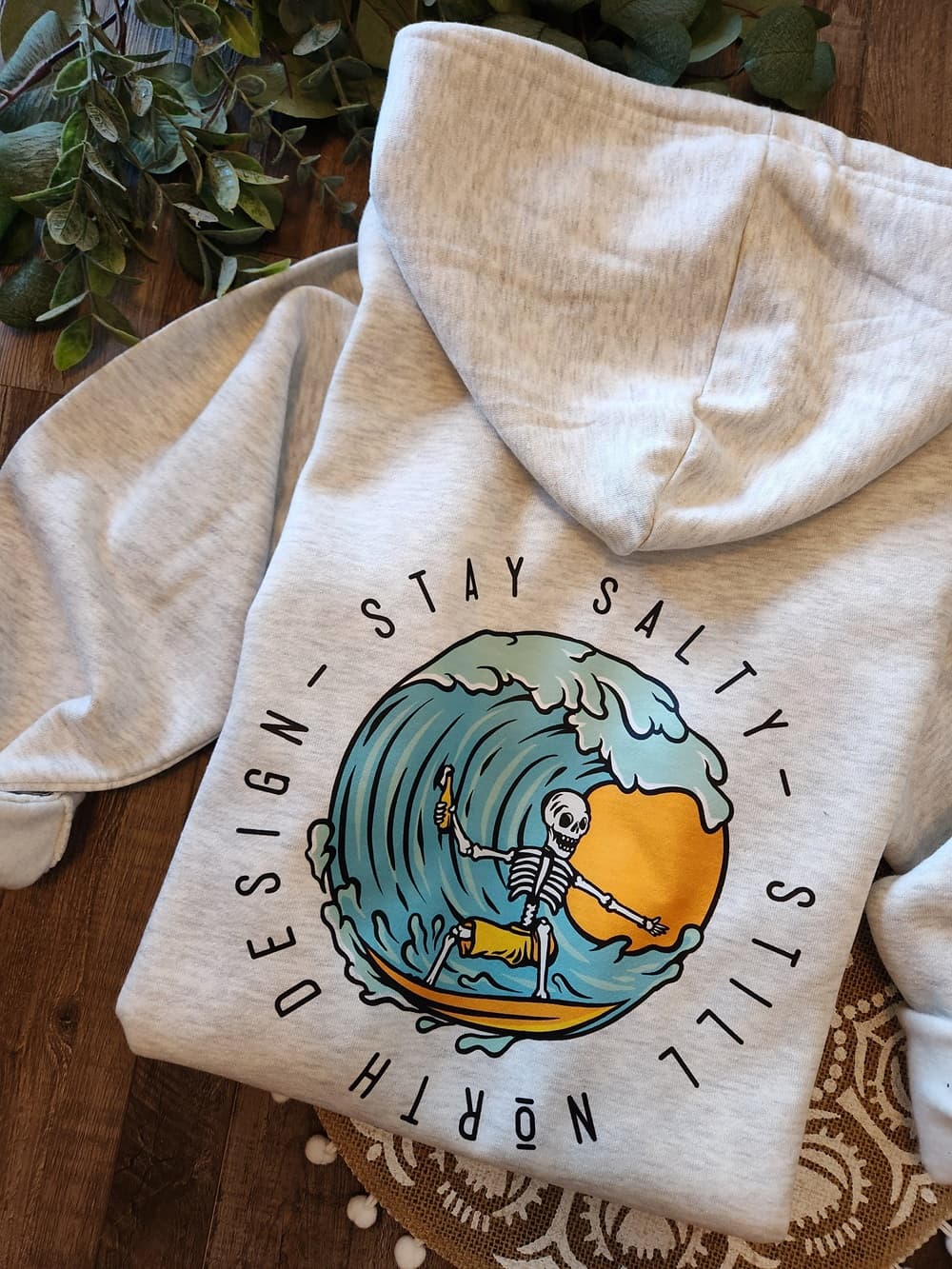 Beige hoodie with a graphic design of a skeleton surfing a wave and text 'Stay Salty' on a wooden surface.