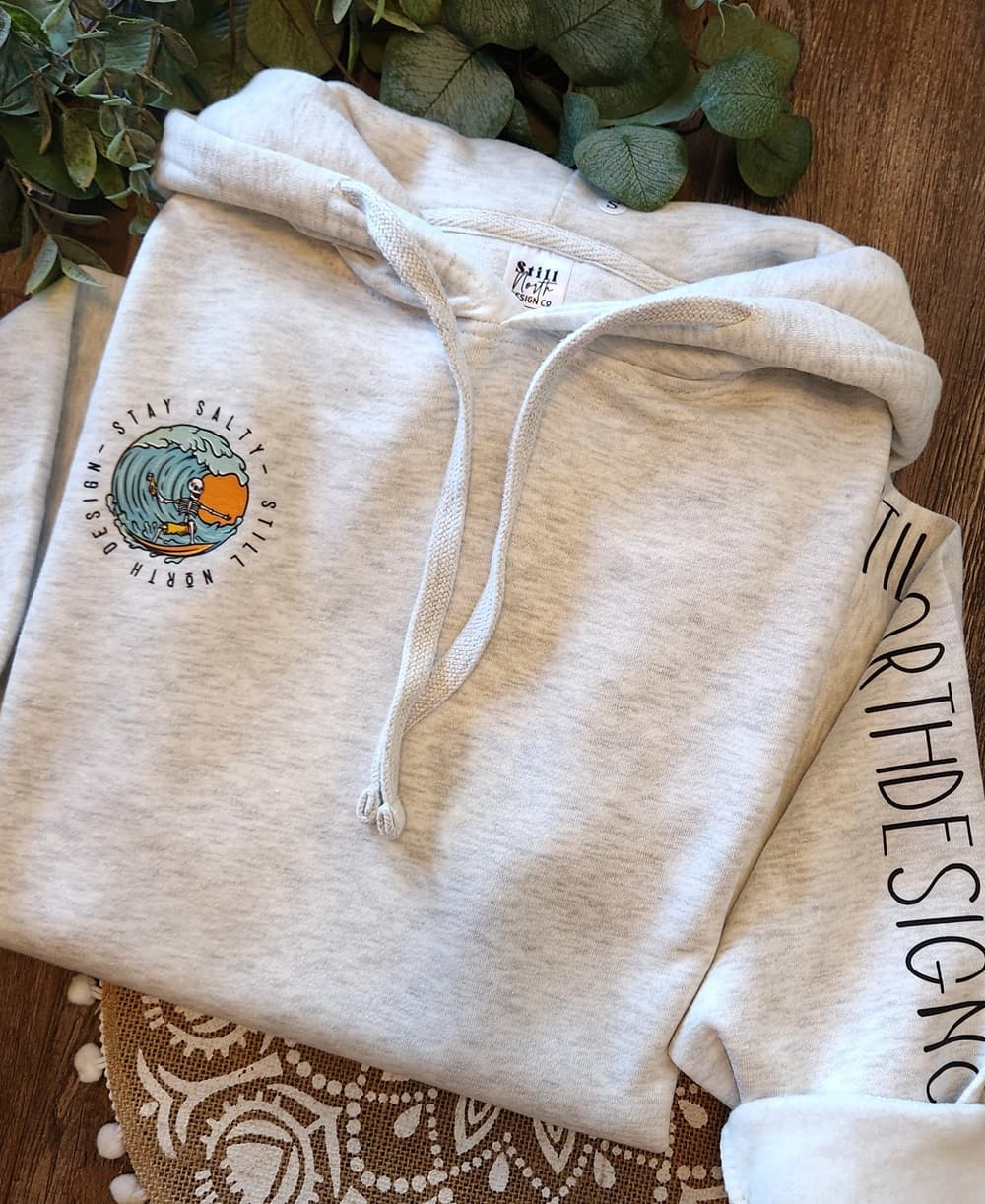 Beige hoodie with a logo on the chest, placed on a wooden surface with greenery in the background.
