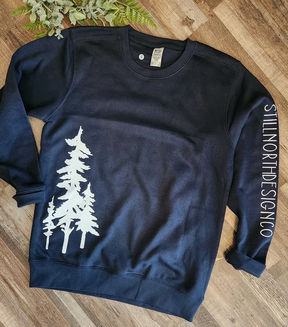 Dark grey sweatshirt with white tree design and 'StillNortnDesignCo' text on a wooden surface.