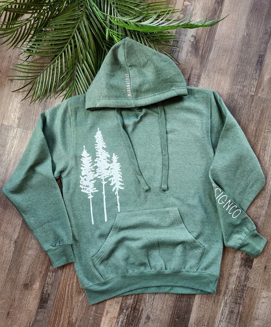 Green hoodie with white tree design on a wooden surface with green leaves.