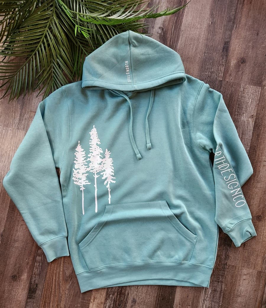 Teal hoodie with white tree design on a wooden surface with green leaves.