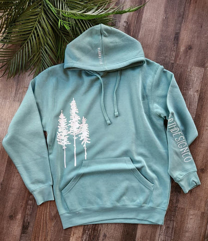 Teal hoodie with white tree design on a wooden surface with green leaves.