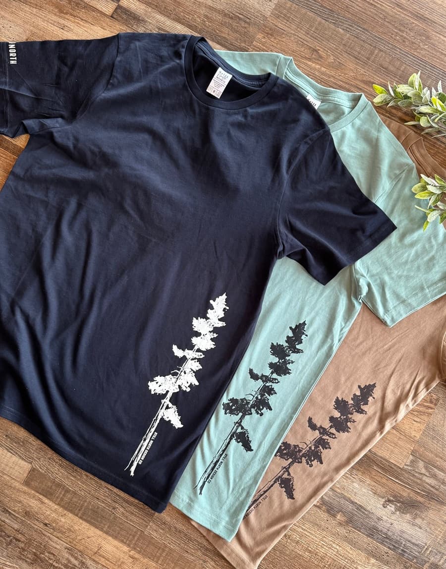 Three t-shirts, one black and one light blue, with tree designs on a wooden surface.