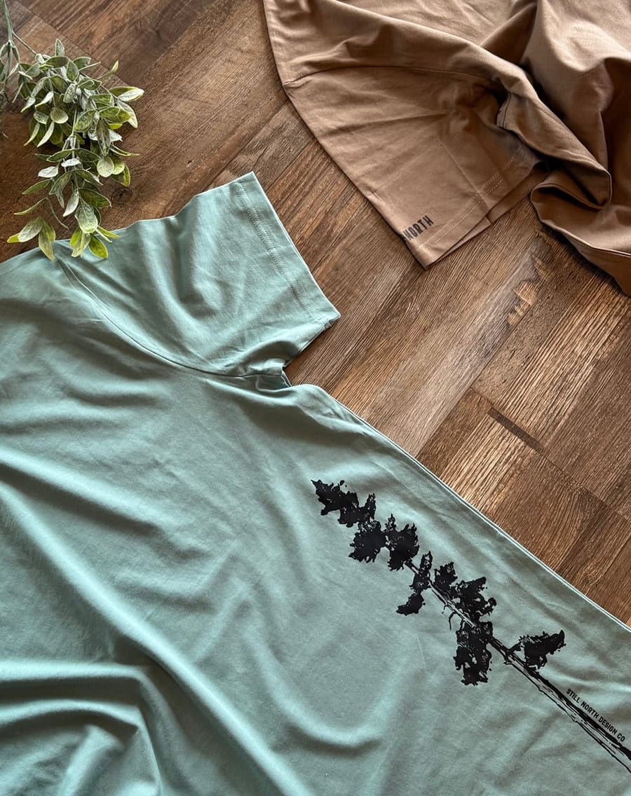 Light blue t-shirt with black leaf design on a wooden floor with a plant and brown fabric.