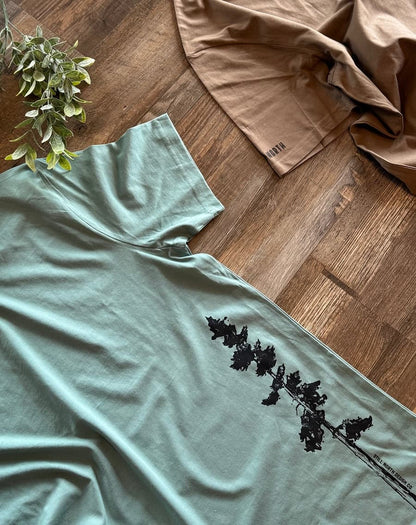 Light blue t-shirt with black leaf design on a wooden floor with a plant and brown fabric.